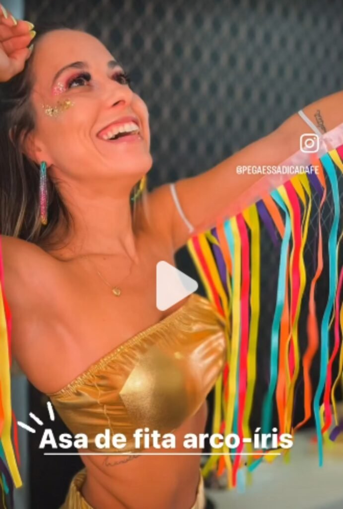 Mulher fantasiada para o carnaval com um top dourado e asas de fita arco-íris, com glitter no rosto.
