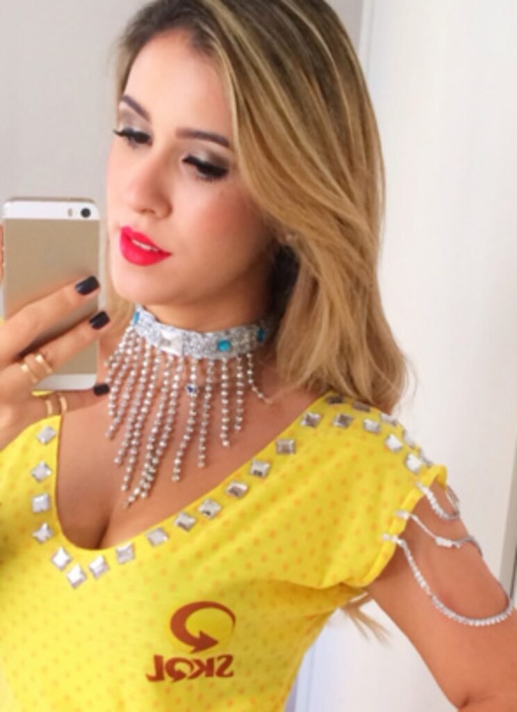 Mulher loira tirando uma selfie no espelho, com uma fantasia de carnaval amarela e acessórios de strass no ombro e no pescoço.