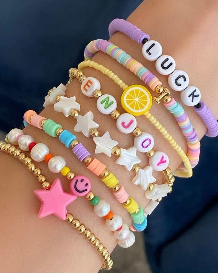 Um pulso com várias pulseiras de miçangas coloridas com as palavras LUCK e ENJOY, com pingentes de estrela, pérola, limão e um emoji sorrindo.